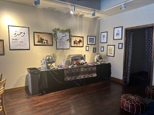 Long table with coffee, tea and food service and appliances in corner with several art pieces hanging on wall